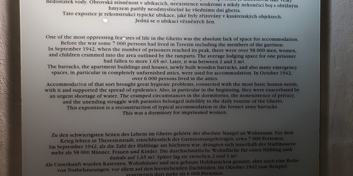 Une des caractéristiques les plus oppressantes de la vie dans le ghetto était le manque absolu d’espace pour se loger. Avant la guerre, environ 7 000 personnes vivaient à Terezín, y compris les membres de la garnison. En septembre 1942, lorsque le nombre de prisonniers atteignit son maximum, plus de 58 000 hommes, femmes et enfants furent entassés dans l’espace délimité par les remparts. La surface moyenne par prisonnier tomba à environ 1,65 m². Par la suite, elle se situa entre 2 et 3 m². Les casernes, les immeubles d’habitation, les maisons, les nouvelles baraques en bois ainsi que de nombreux espaces d’urgence, notamment des greniers non aménagés, furent utilisés comme logements. En octobre 1942, plus de 6 000 personnes vivaient dans les greniers. Ces conditions entraînèrent de graves problèmes d’hygiène liés aux besoins humains les plus élémentaires et favorisèrent la propagation des épidémies. Au début notamment, elles furent aggravées par une pénurie d’eau. La promiscuité extrême dans les dortoirs, l’absence totale d’intimité et la lutte permanente contre les parasites faisaient partie du quotidien du ghetto. Cette exposition est une reconstitution des conditions typiques de logement dans les anciennes casernes. Il s’agit d’un dortoir pour femmes prisonnières.